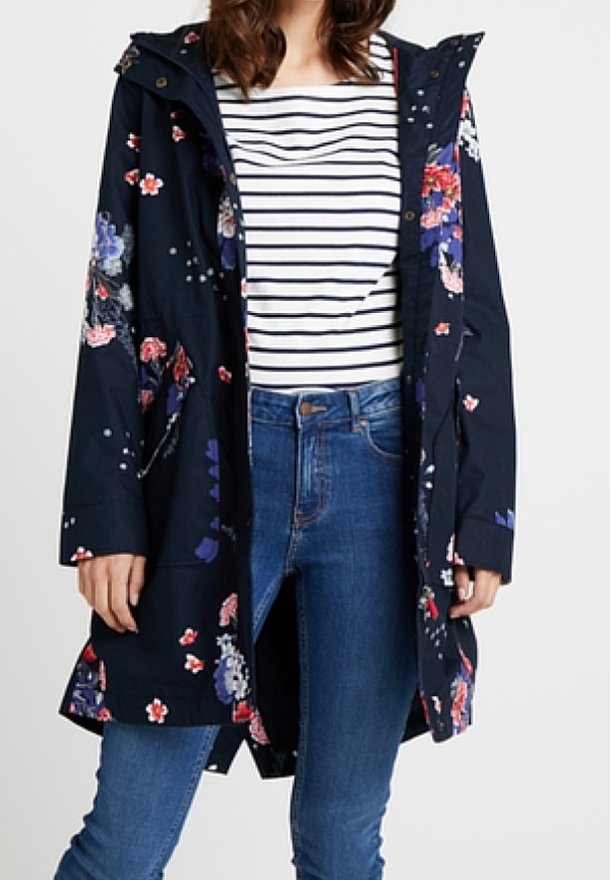 Marineblå blomstret regnfrakke med hætte, prydet med pink og hvide blomster, en knaplukning foran og lommer, båret over en stribet top og jeans.