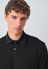 Juodas polo marškinėlis su languotu žaliai juodu apykakle, dviejų mygtukų eilute ir mažuoju karūnos logotipu ant krūtinės. Lygus tekstūra.