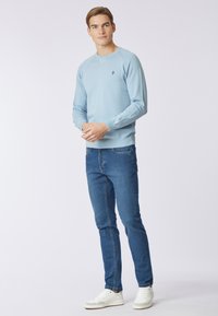 Maglione azzurro chiaro a girocollo in tessuto morbido, abbinato a jeans blu scuro. Il modello sta in piedi in modo casual, indossando sneakers bianche. Design minimale.