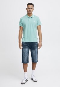 Menta polo majica s bijelim detaljima, uparena s plavim trapericama. Model nosi bijele čarape i Adidas tenisice. Jednostavan, ležeran outfit.