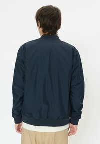 Personne portant une veste bleu marine, un pantalon beige et une chemise blanche, vue de dos sur un fond blanc uni.