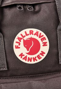 Στρογγυλή λευκή ετικέτα με κόκκινο στυλιζαρισμένο λογότυπο αλεπού και κείμενο "Fjällräven Kånken" σε ύφασμα σκούρου καφέ σακιδίου.