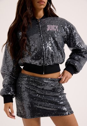 Naine kannab säravat musta litritega lühikest jakki teksti "JUICY" ja sobivat litritega miniseelikut, käsi toetub seeliku vöökohale.