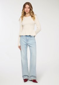 Maglia bianca a costine a maniche lunghe con orlo svasato, abbinata a jeans a gamba larga azzurri e scarpe rosse con punta appuntita. Texture uniforme in tutta la superficie.