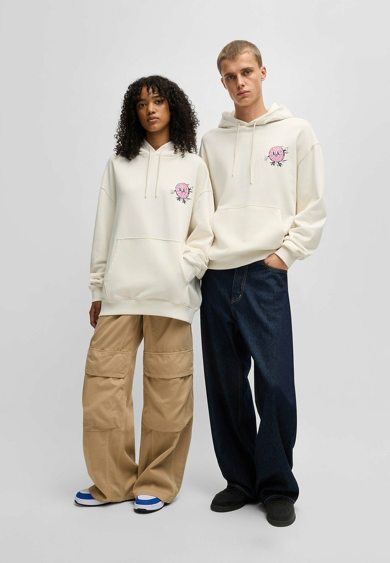 Hoodie crème avec une poche frontale et un motif graphique rose sur la poitrine. Le mannequin porte un pantalon cargo beige et des chaussures avec des accents bleus.