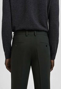 Tył osoby ubranej w ciemnozielone, dopasowane spodnie z zapinanymi na guziki tylnymi kieszeniami oraz wciśnięty w spodnie ciemnoszary sweter z długim rękawem.