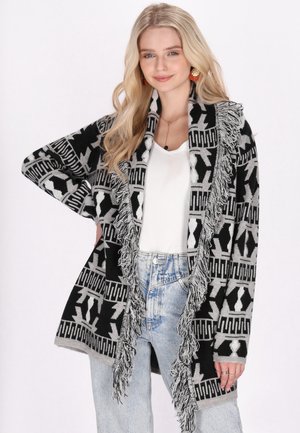 Cardigan a righe nere e grigie con frange lungo i bordi, vestibilità morbida, texture soffice, indossato sopra una maglietta bianca e abbinato a jeans a vita alta.