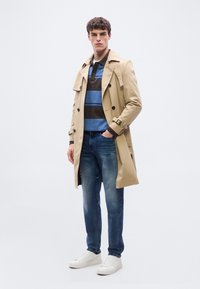 Άνδρας που φοράει μπεζ παλτό τρεντσκότ, μπλε και καφέ ριγέ πόλο, μπλε τζιν και άσπρα αθλητικά παπούτσια, στέκεται με το ένα χέρι στην τσέπη.