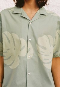 Jasnozielona koszula na krótki rękawek zapinana na guziki, ozdobiona dużymi, jasnoniebieskimi nadrukami tropikalnych liści, noszona przez osobę z kręconymi, ciemnymi włosami.