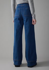 Wide-leg blue denim pants with a high waist, featuring back pockets and contrasting red stitching along the seams. Smooth texture.