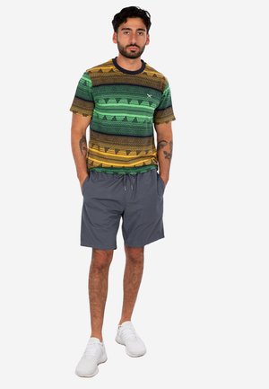 Mann steht mit den Händen in den Taschen, trägt ein mehrfarbig gestreiftes T-Shirt, graue Shorts, weiße Turnschuhe und hat Tätowierungen an den Armen.