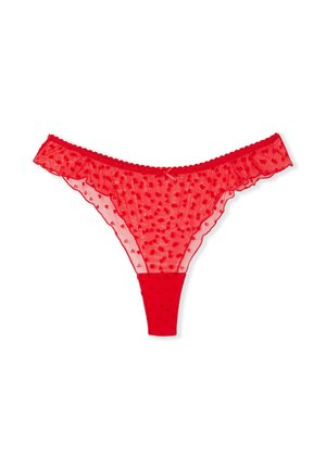 Femme portant un haut blanc ajusté à manches courtes et une lingerie rouge semi-transparente à motifs de cœurs, debout devant un fond blanc uni.