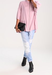 Helles rosa, kurzärmeliges, mit Reißverschluss versehenes Shirt, kombiniert mit patchwork-mäßigen, hellblauen und weißen Jeans. Hält eine schwarze Handtasche und trägt schwarze Stiefeletten mit Absatz.