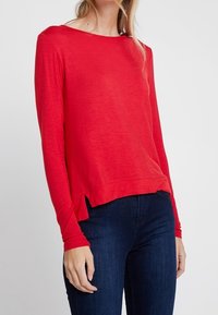 Rotes langärmliges Strickoberteil mit Bootsausschnitt, ausgestattet mit seitlichen Schlitzen am Saum, kombiniert mit dunkelblauen Jeans. Weiche Textur und leichtes Material.
