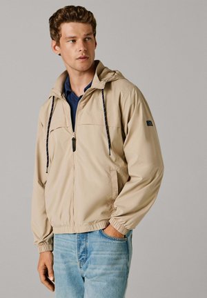 Beige letvægtsjakke med hætte, justerbare snore, lynlås, sidelommer, elastiske manchetter, båret over en navy skjorte og jeans.