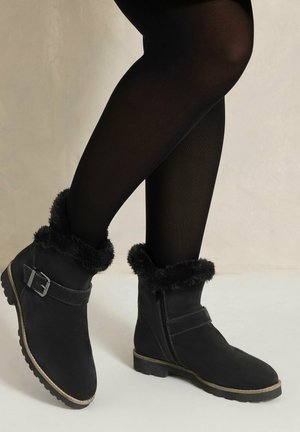 Bottines noires avec bordure en fourrure et détail boucle portées avec des collants noirs sur un fond clair.