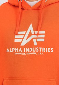 Sudadera naranja de mezcla de algodón, con texto impreso en blanco y un logo. Incluye capucha con cordón y bolsillo frontal.