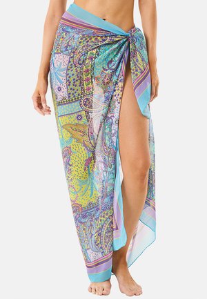 Donna che indossa un sarong colorato e decorato, legato in vita, con una spacco alto che mostra una gamba nuda, in piedi a piedi nudi su sfondo bianco.