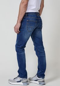 Jeans de mezclilla azul oscuro con un diseño ajustado, que cuentan con dos bolsillos traseros, detalles de costura sutiles y una textura suave. Combinados con zapatillas gris.