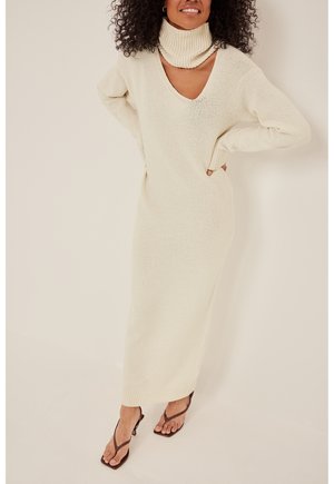 Robe pull longue en crème avec un col en V et un col haut. Présente des poignets côtelés et une coupe décontractée, fabriquée en tissu tricoté doux.