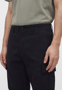 Man wearing black cargo shorts and olive green t-shirt, hand resting near flap pocket on right thigh.