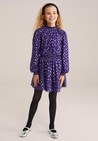 Ljubičasta haljina sa smokanom strukom i dugim rukavima, s bijelim apstraktnim uzorcima. Uparena s crnim tajicama i srebrnim cipelama.