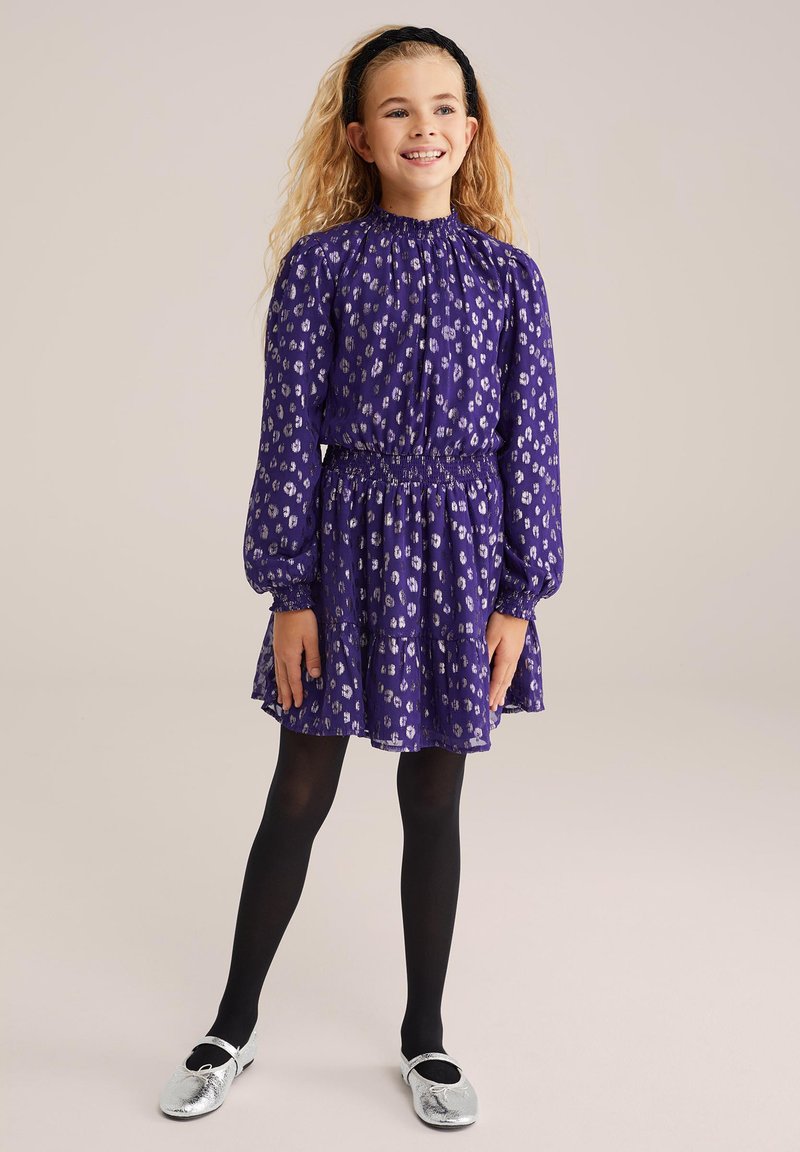 Ljubičasta haljina sa smokanom strukom i dugim rukavima, s bijelim apstraktnim uzorcima. Uparena s crnim tajicama i srebrnim cipelama.