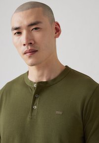 Zeleni dolgi rokav henley majica iz mehkega materiala, z tremi gumbi na ovratniku in majhnim logotipom na prsih.