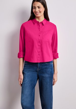 Nainen hymyilee ja pitää päällään kirkkaanpunaista napillista paitaa, jossa on käärityt hihat, sekä sinisiä farkkuja, seisten vaaleaa taustaa vasten.