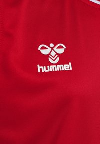Czerwony materiał sportowy z wyraźnym białym logo "hummel" z elementami pszczoły i chevronu. Gładka tekstura, z widocznymi detalami szycia.