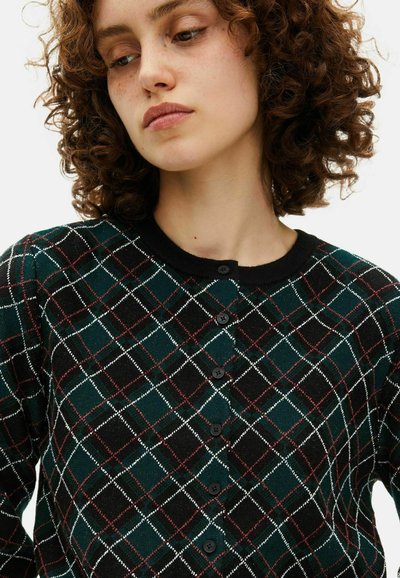 Donna con i capelli ricci che indossa un cardigan a scacchi con bottoni di colore verde scuro, nero, bianco e rosso, che guarda verso il basso e leggermente di lato.