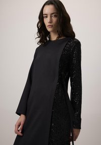 Vestido negro con un lado de lentejuelas texturizadas y un panel de tela suave, mangas largas y un detalle atado en la cintura.