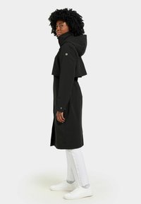Manteau noir imperméable au design court, avec capuche et boutons pression. Porté avec un pantalon blanc et des baskets, présentant une texture lisse.