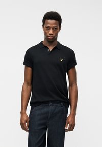 Νέος άνδρας που φοράει μαύρο πόλο με κοντά μανίκια και χρυσό έμβλημα πουλιού, καθώς και σκούρο τζιν παντελόνι, στέκεται μπροστά σε απλό λευκό φόντο.