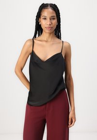 Zwarte satijnen camisole met een V-hals en dunne bandjes, gedragen met bordeauxrode wijduitlopende broek. Soepele textuur, zachtvallend over het lichaam.