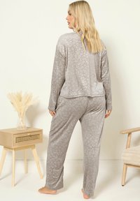 Kvinna med långt blont hår står barfota, vänd bort, iförd en mjuk grå leopardmönstrad pyjamas i ett minimalistiskt rum med trämöbler.