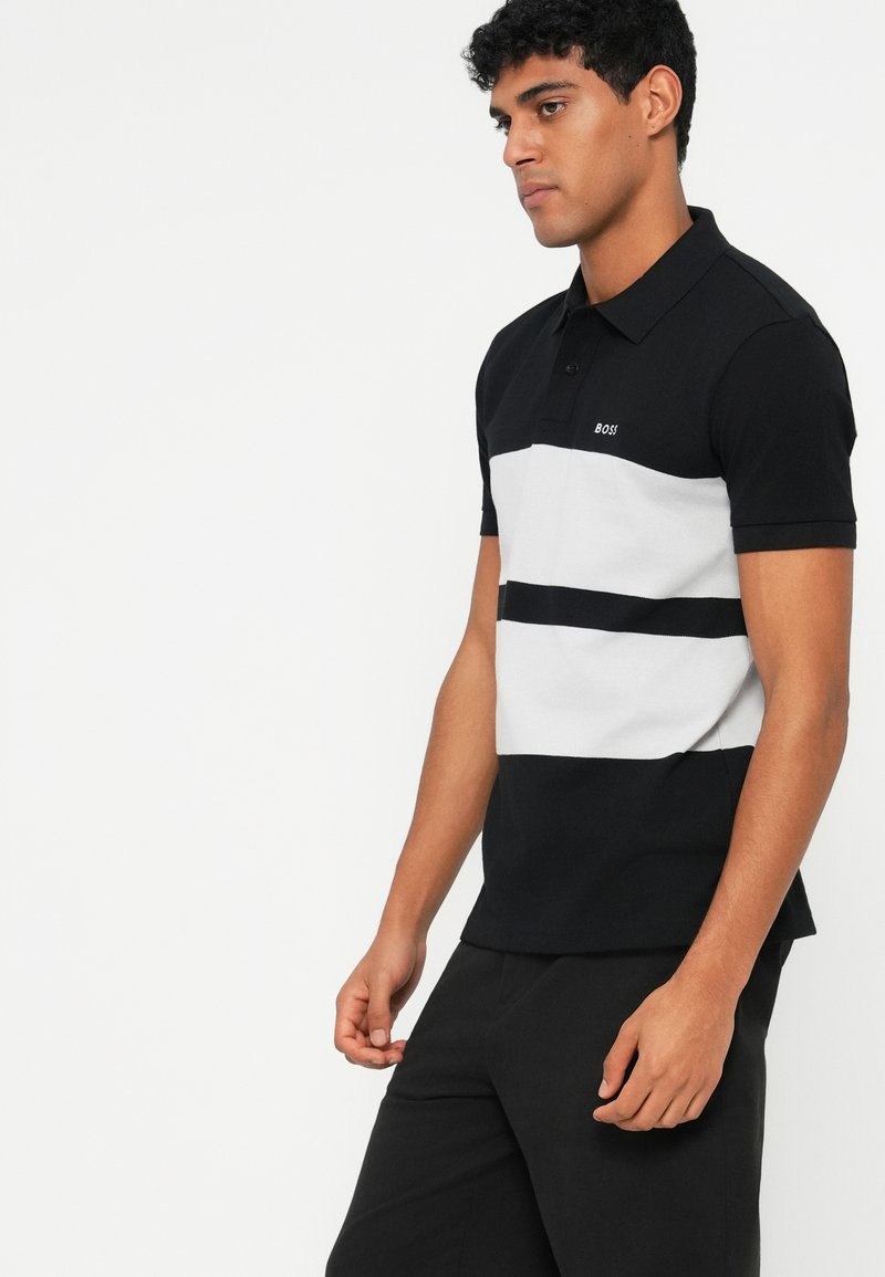 Camisa polo a rayas negras y grises con un collar clásico, que presenta un pequeño logotipo "BOSS" en el pecho, combinada con pantalones negros.