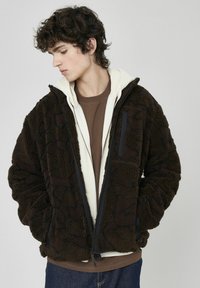 Veste en polaire marron avec un extérieur texturé et à motif. Elle dispose d'une fermeture éclair à l'avant et d'un design à double couche avec une couche intérieure de couleur claire.