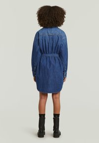 Τζιν φόρεμα σε σκούρο μπλε, με στενή μέση, μακριά μανίκια, κουμπιά στους καρπούς και στρογγυλή άκρη. Φορεμένο με μαύρες μπότες μέχρι τον αστράγαλο.