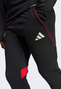 Pantaloni sportivi neri realizzati in tessuto elasticizzato, con un pannello laterale rosso e un logo riflettente bianco sulla coscia. Tessuto liscio.