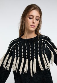 Schwarzer gestrickter Pullover mit strukturiertem Stoff, weißen Quastenakzenten und einem runden Ausschnitt mit kontrastierenden vertikalen Streifen.