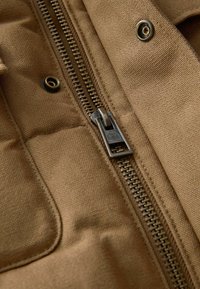 Pruun kangas, millel on tikitud tekstuur, koos metallist tõmbluku, messingist komponentide ja tugevdatud õmblustega.