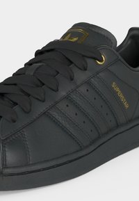 Μαύρο αθλητικό παπούτσι Adidas Superstar με χρυσό λογότυπο, χρυσή τρύπα για τα κορδόνια, μαύρα κορδόνια και τη λέξη "SUPERSTAR" στην πλευρά, εμφανιζόμενο από κοντινή γωνία.
