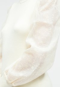 Valkoinen ribbineuloksinen toppi, jossa läpikuultavat kirjotut pussihihat kukkakuvioin. Pehmeä rakenne ja hieman vartalonmyötäinen siluetti.