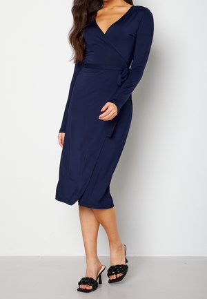 Femme portant une robe portefeuille bleu marine à manches longues avec un lien sur le côté et des sandales noires tressées à talons hauts et bout ouvert.