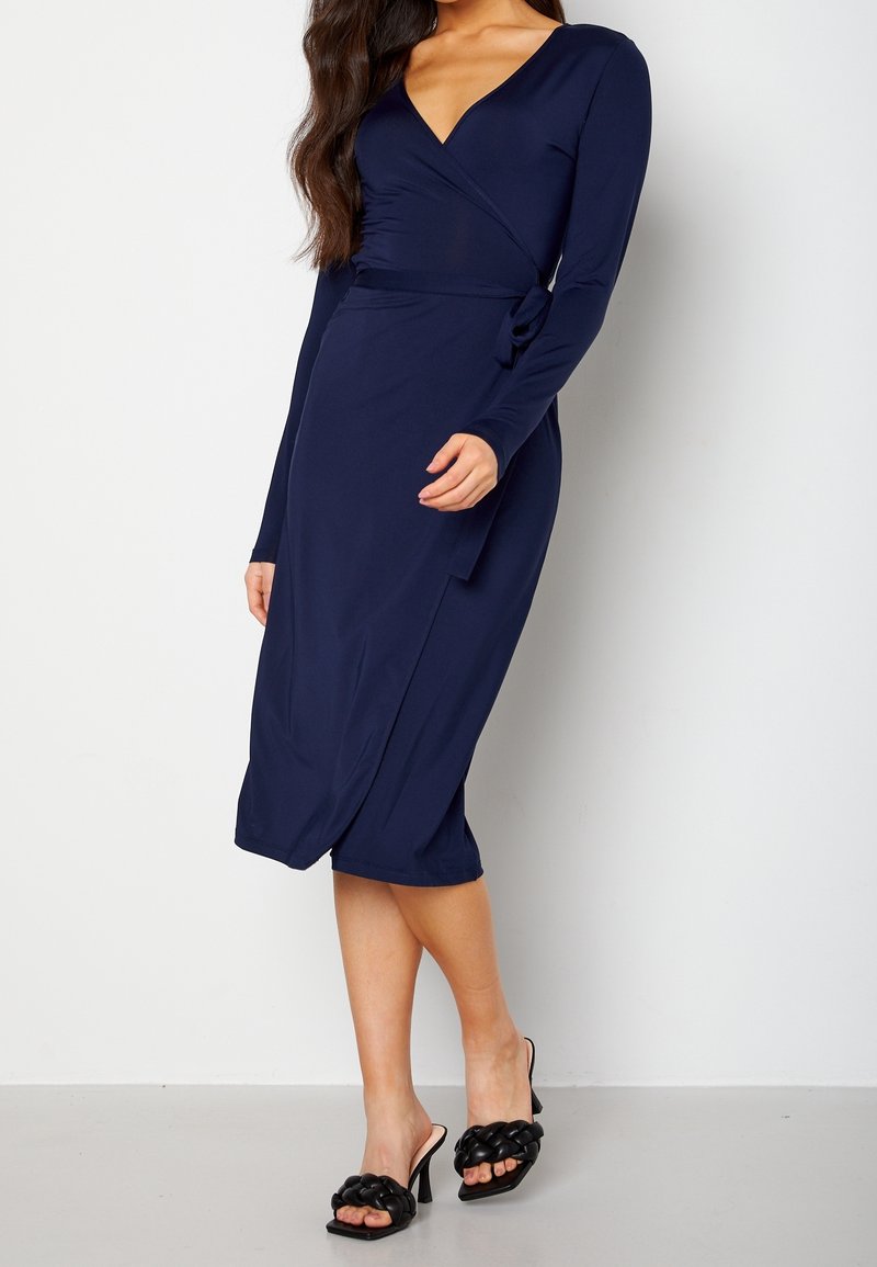 Femme portant une robe portefeuille bleu marine à manches longues avec un lien sur le côté et des sandales noires tressées à talons hauts et bout ouvert.