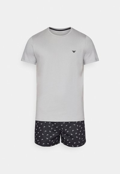 Graues Baumwoll-T-Shirt mit Rundhalsausschnitt und kurzen Ärmeln, kombiniert mit schwarzen Badehosen, die ein weißes wiederholtes Logo-Muster aufweisen.