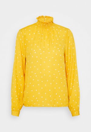 Blusa gialla con maniche lunghe, colletto alto e arricciato, e pois bianchi. Realizzata in un tessuto leggero con una superficie testurizzata.