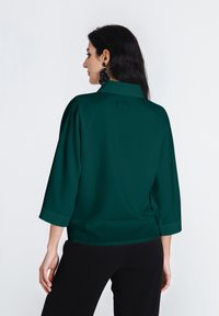 Türki, 3/4 pikkusega blouse, millel on krae, sile tekstuur ja lahtine lõige. Rikkalikuks mustade kõrvarõngastega, kombineeritud mustade pükstega.
