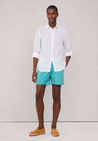 Camicia bianca di lino, maniche arrotolate, pantaloni corti turchesi con motivo, e espadrillas marroni. Design semplice e tessuto leggero.